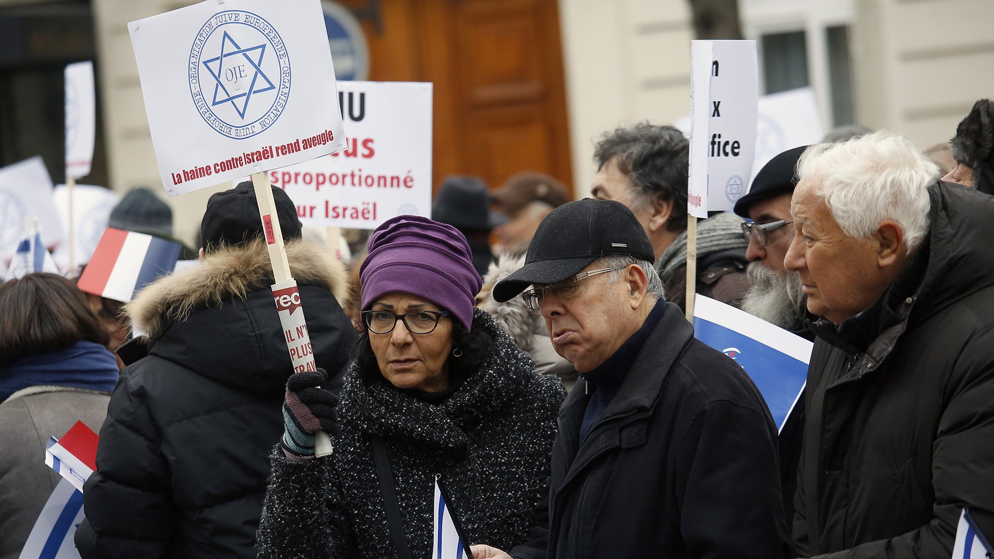 در این تصویر، گروهی از افراد در حال اعتراض هستند. آن‌ها پلاکاردها و تابلوهای مختلفی در دست دارند. برخی از افراد لباس‌های گرم به تن دارند و در میان آن‌ها یک زن با عینک دیده می‌شود. تابلوها شامل پیام‌هایی مرتبط با اسرائیل هستند و برخی از افراد به سمت دوربین نگاه می‌کنند. فضای تجمع در یک مکان عمومی به نظر می‌رسد.