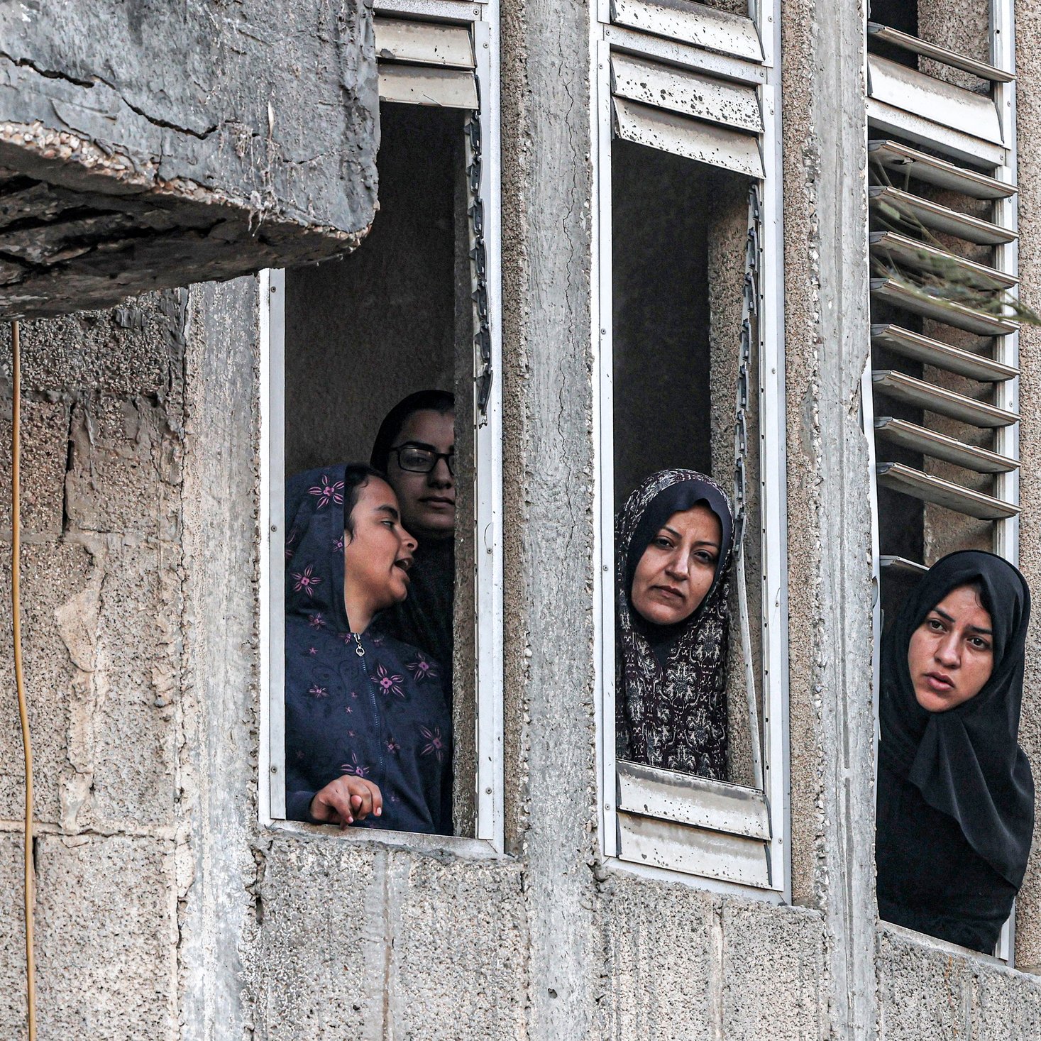 L'image montre plusieurs femmes regardant par des fenêtres d'un bâtiment. Elles semblent attentives et curieuses, certaines portant des hijabs. Le mur extérieur du bâtiment est en béton, et on peut apercevoir des fenêtres aux volets cassés. L'ambiance paraît chargée d'émotions, mais on ne peut pas déterminer le contexte exact de la scène.
