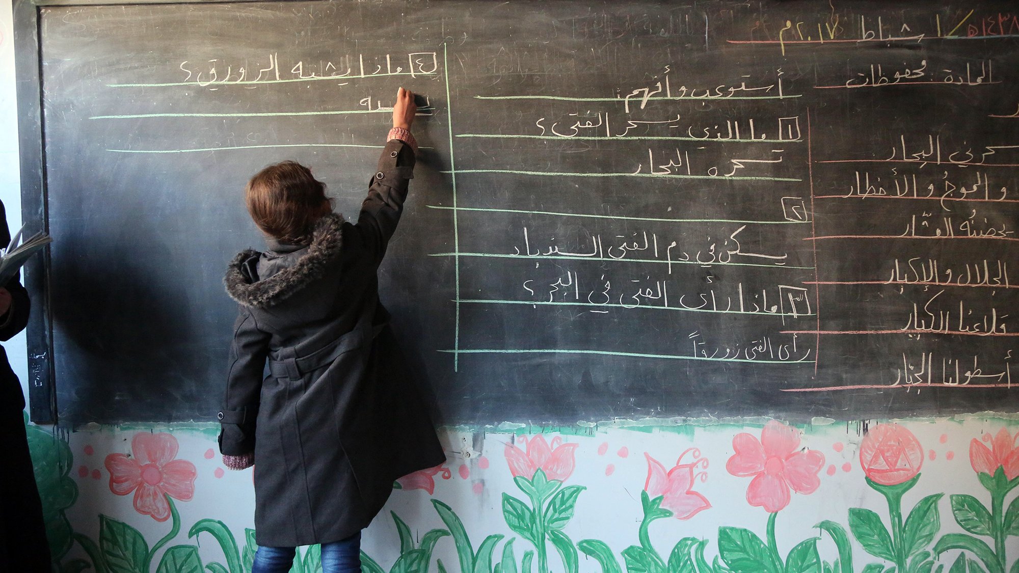 L'image montre une salle de classe avec un tableau noir. Une élève, de dos, écrit quelque chose au tableau avec une craie blanche. Elle porte un manteau noir. Le tableau est divisé en plusieurs sections avec des inscriptions en arabe. En bas du tableau, on peut voir des dessins de fleurs colorées qui décorent le mur. L'ambiance générale semble être celle d'un environnement scolaire.