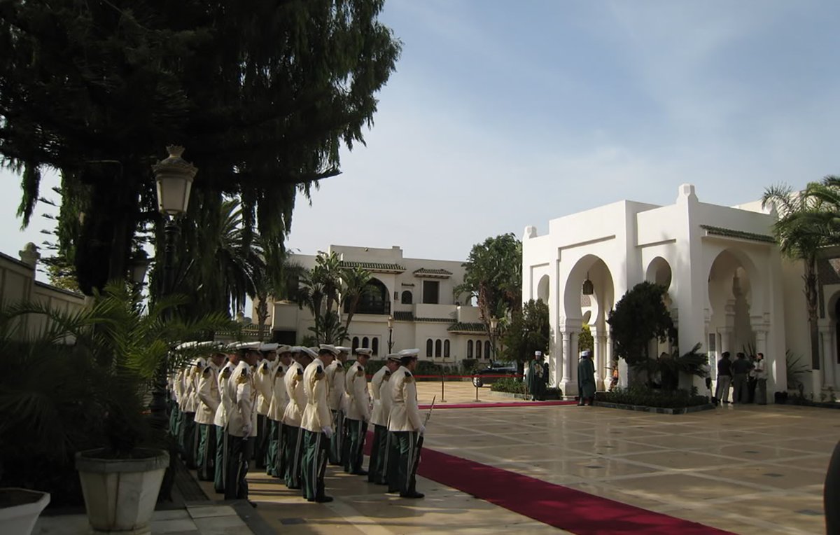 L'image montre une scène extérieure d'un bâtiment élégant, probablement un palais ou un hôtel de luxe. On peut voir une allée centrale avec un tapis rouge, entourée de verdure et de palmiers. À gauche, un groupe de personnes en uniformes formés, représentant une sorte de garde ou de cérémonie. En arrière-plan, on aperçoit un bâtiment majestueux avec des arches et des colonnes, suggérant une architecture raffinée. Le ciel est clair, ce qui ajoute à l'atmosphère solennelle de la scène.
