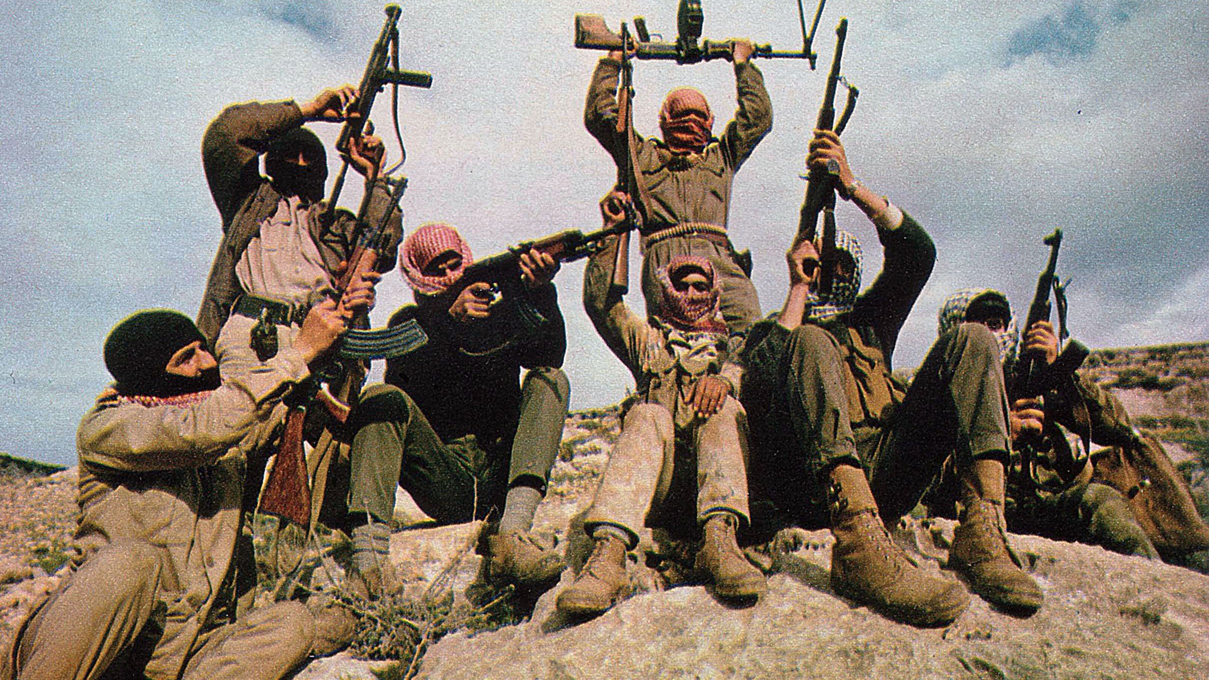 L'image montre un groupe de personnes armées, portant des masques ou des couvre-visages, prenant une pose militante sur un rocher. Ils tiennent des armes à feu en l'air et semblent être en pleine démonstration de force. Le paysage est naturel, probablement en extérieur, avec un ciel nuageux en arrière-plan. L'ensemble de la scène véhicule une atmosphère de tension ou de préparation pour une action.