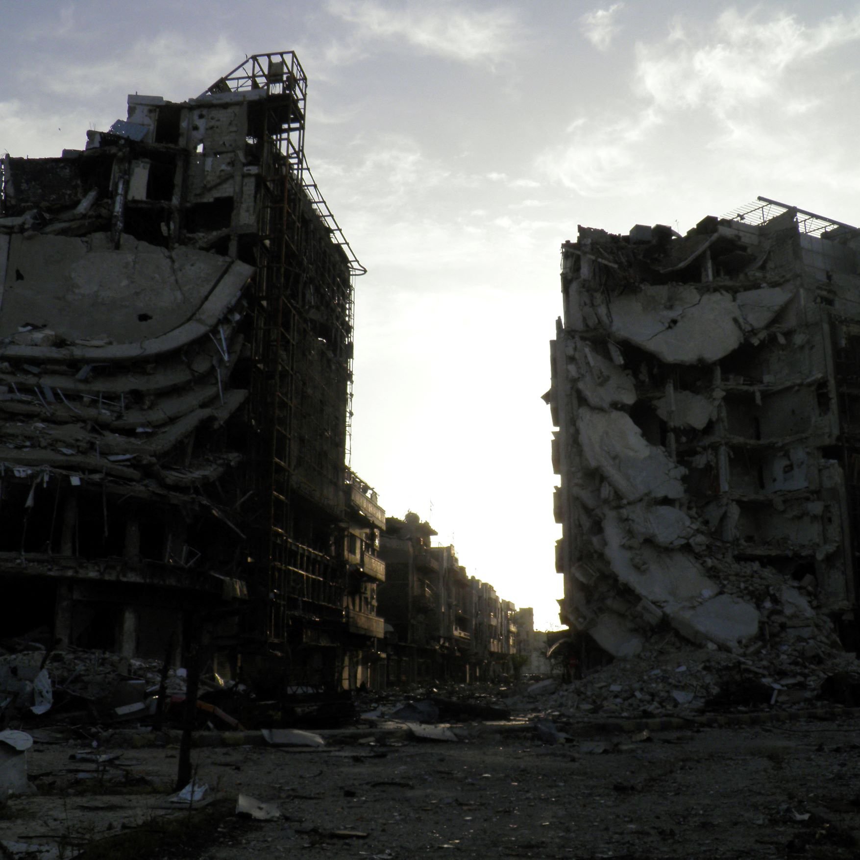تظهر الصورة عمارتين محطمتين في منطقة يبدو أنها تعرضت لتدمير واسع. المباني مشوهة، حيث تظهر الجدران المنهارة والأسطح المدمرة، مما يعكس آثار الدمار. السماء في الخلفية تبدو ملبدة، مما يضيف شعورًا بالخيبة والخراب. المنطقة المحيطة يبدو أنها مهجورة، مما يعكس الأثر العميق للصراعات أو الكوارث التي أدت إلى هذا الوضع.