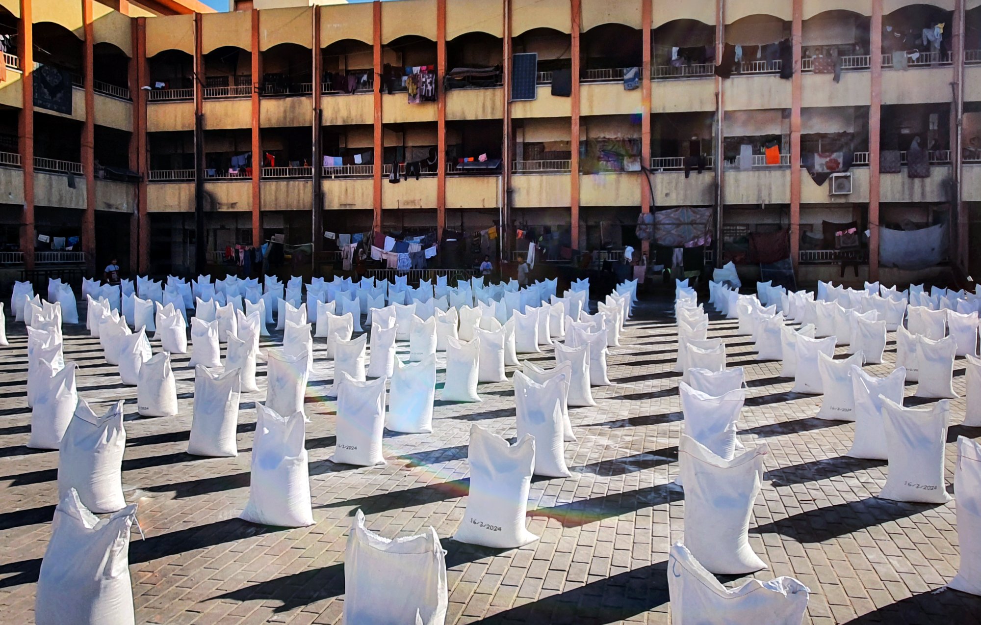L'immagine mostra un'area all'aperto con molti sacchi bianchi disposti in file ordinate. Sullo sfondo si vede un edificio di più piani, con diversi balconi. Alcuni panni sono appesi ad asciugare tra i balconi. La scena è illuminata dalla luce del sole, creando un'atmosfera vivace e attiva. Il pavimento è costituito da mattonelle in cui si possono notare i segni del tempo. L'insieme suggerisce un contesto urbano con attività legate alla distribuzione di beni.