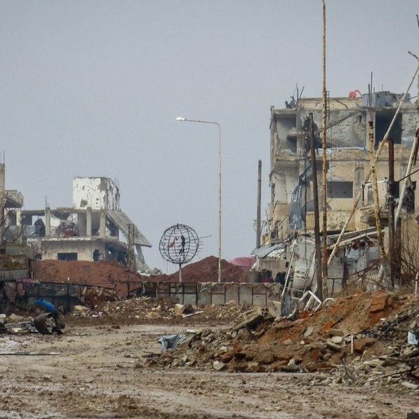 L'image montre une scène de destruction urbaine. On peut voir des bâtiments en ruines, avec des murs effondrés et des structures endommagées. Le sol est recouvert de débris, et l'atmosphère semble sombre et désolée. Il y a des poteaux électriques dans l'arrière-plan, et un éclairage public est visible au loin, mais tout cela contribue à une ambiance de désolation. La végétation est rare, et l'ensemble dégage une impression de lieu abandonné et ravagé.