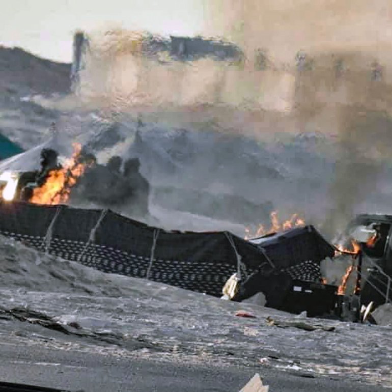 L'image semble représenter une scène de conflit ou de guerre, avec des flammes visibles et de la fumée s'élevant dans l'air. On peut distinguer des structures endommagées et une ambiance chaotique. Le sol est recouvert de débris, et des véhicules militaires ou des équipements peuvent être présents. L'environnement semble aride, suggérant un paysage désertique.