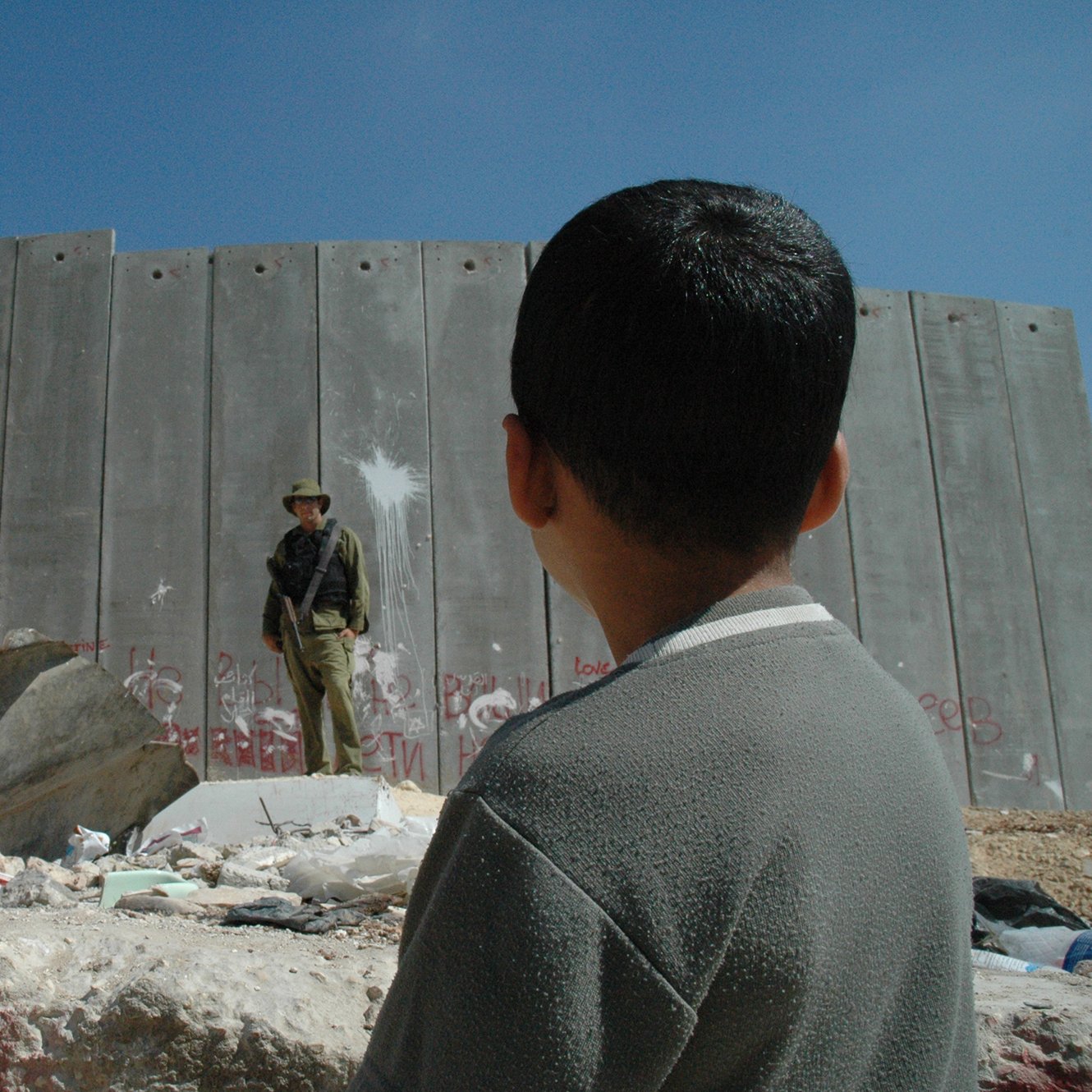 در این تصویر، پسر بچه‌ای به سمت یک دیوار بلند و بتنی نگاه می‌کند. دیوار دارای آثار graffiti و علامت‌های مختلفی است. در مقابل او، یک نظامی ایستاده است که لباس نظامی به تن دارد و به نظر می‌رسد در حال نظارت است. محیط اطراف شامل سنگ‌ها و زباله‌های پراکنده است و آسمان آبی و روشنی بالای سر آنها دیده می‌شود. احساس تصویر می‌تواند تنش و جدایی را به خوبی نشان دهد.