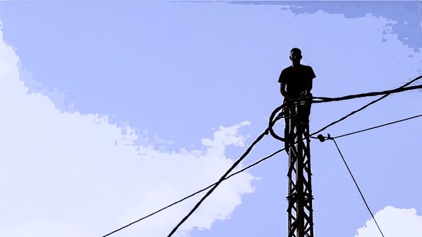 Dans cette image, on peut voir un homme debout sur un pylône, entouré de câbles. Le ciel en arrière-plan est bleu avec quelques nuages. La silhouette de l'homme est mise en avant par le contraste avec le ciel. L'atmosphère semble à la fois audacieuse et impressionnante, soulignant une certaine prise de risque.