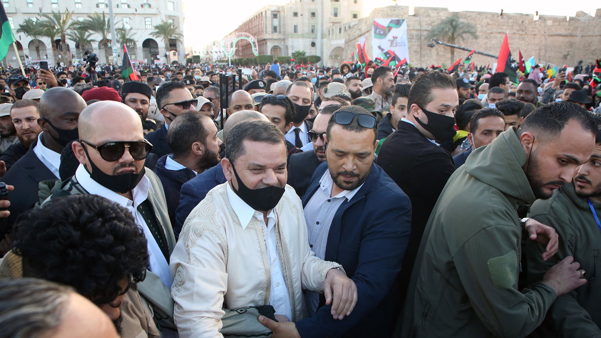 در این تصویر، جمعیتی از مردم در یک رویداد یا مراسم خیابانی دیده می‌شود. در مرکز تصویر، مردی در لباس سنتی روشن در حال احاطه شدن توسط چند نفر دیگر است که بعضی از آن‌ها ماسک زده‌اند. به نظر می‌رسد که او مورد توجه و استقبال زیادی قرار گرفته و در حال تعامل با دیگران است. فضای اطراف پر از افرادی است که در حال تماشا یا شرکت در این رویداد هستند و پرچم‌هایی در دست دارند. در زمینه، ساختمان‌ها و فضاهای عمومی نیز مشخص است.