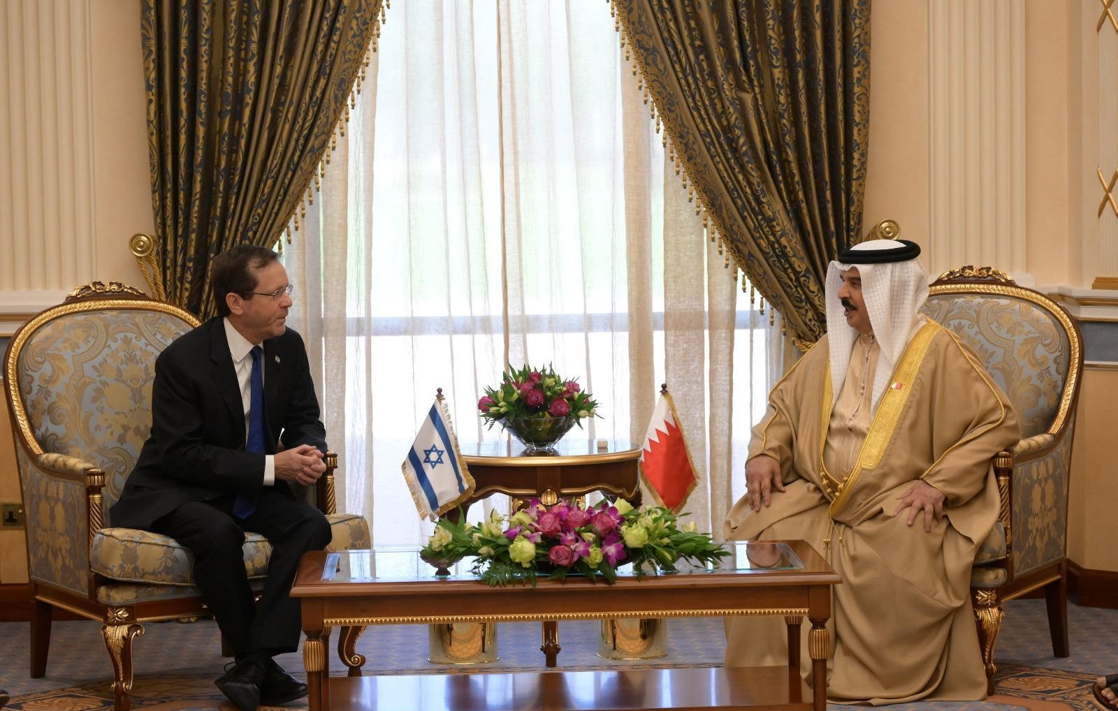 Two men in formal attire sit across from each other in a richly decorated room, discussing.