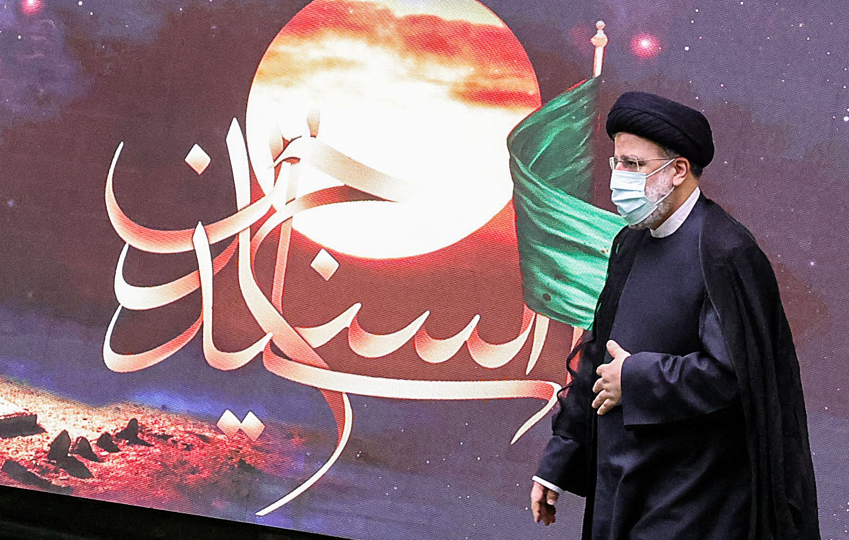 در این تصویر، یک مرد در لباس رسمی و با عمامه مشکی در حال راه رفتن است. او ماسک به صورت دارد و پشت او تصاویری از یک پس‌زمینه هنری با رنگ‌های گرم و نمادهایی مربوط به تاریخ یا فرهنگ دیده می‌شود، از جمله پرچم سبز. متن خوشنویسی شده‌ای در تصویر وجود دارد که به زبان فارسی نوشته شده است. این تصویر ممکن است به یک مراسم مذهبی یا ملی مرتبط باشد.