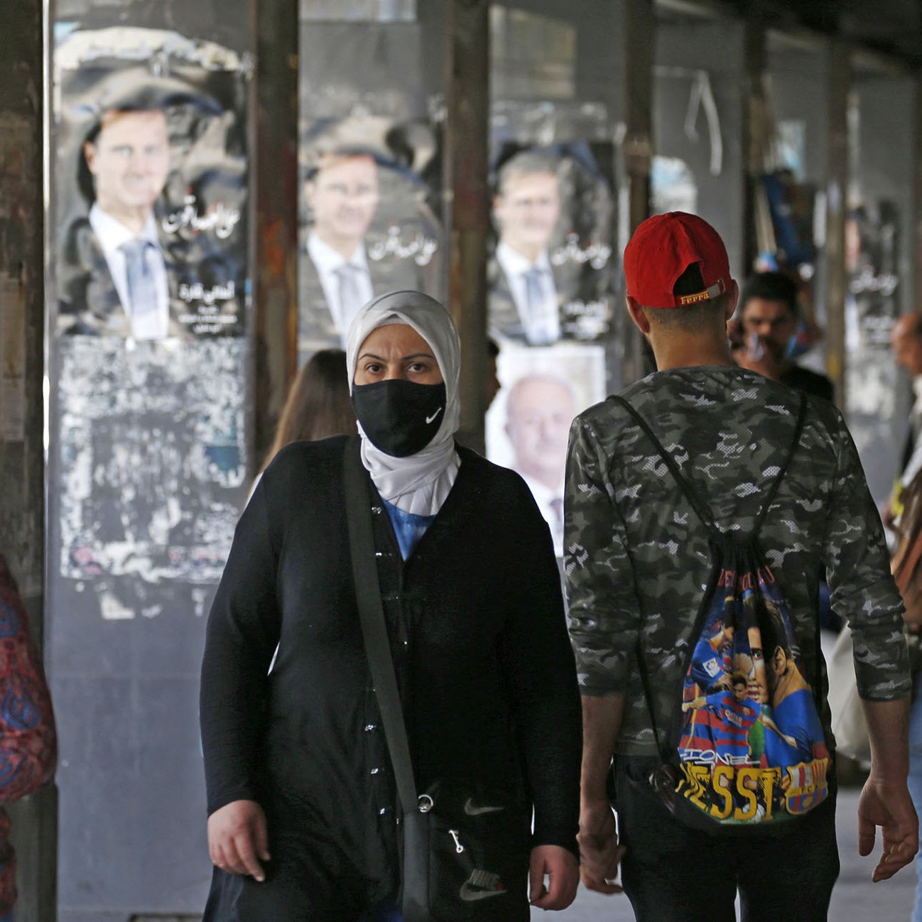 Nell'immagine si vedono diverse persone in un'area urbane, probabilmente un mercato o una strada affollata. Alcuni passanti indossano mascherine, mentre altri non ne hanno. Sullo sfondo, ci sono manifesti con l'immagine di un uomo, presumibilmente un leader politico, appesi alle pareti. L'atmosfera sembra quella di un luogo vivo e multiculturale, con persone di diverse età e stili che si muovono nella zona.