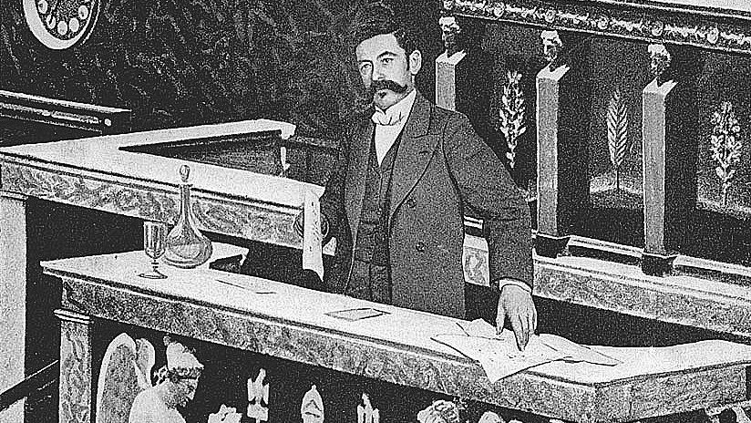 The image depicts a man standing at a podium in what appears to be a legislative or parliamentary setting. He is dressed in a formal suit and has a mustache. The podium is richly decorated, with sculptures of angelic figures and ornate designs. There seem to be documents and a glass on the podium, indicating a formal presentation or discussion taking place. The background suggests an elegant and historical architecture, typical of governmental buildings.