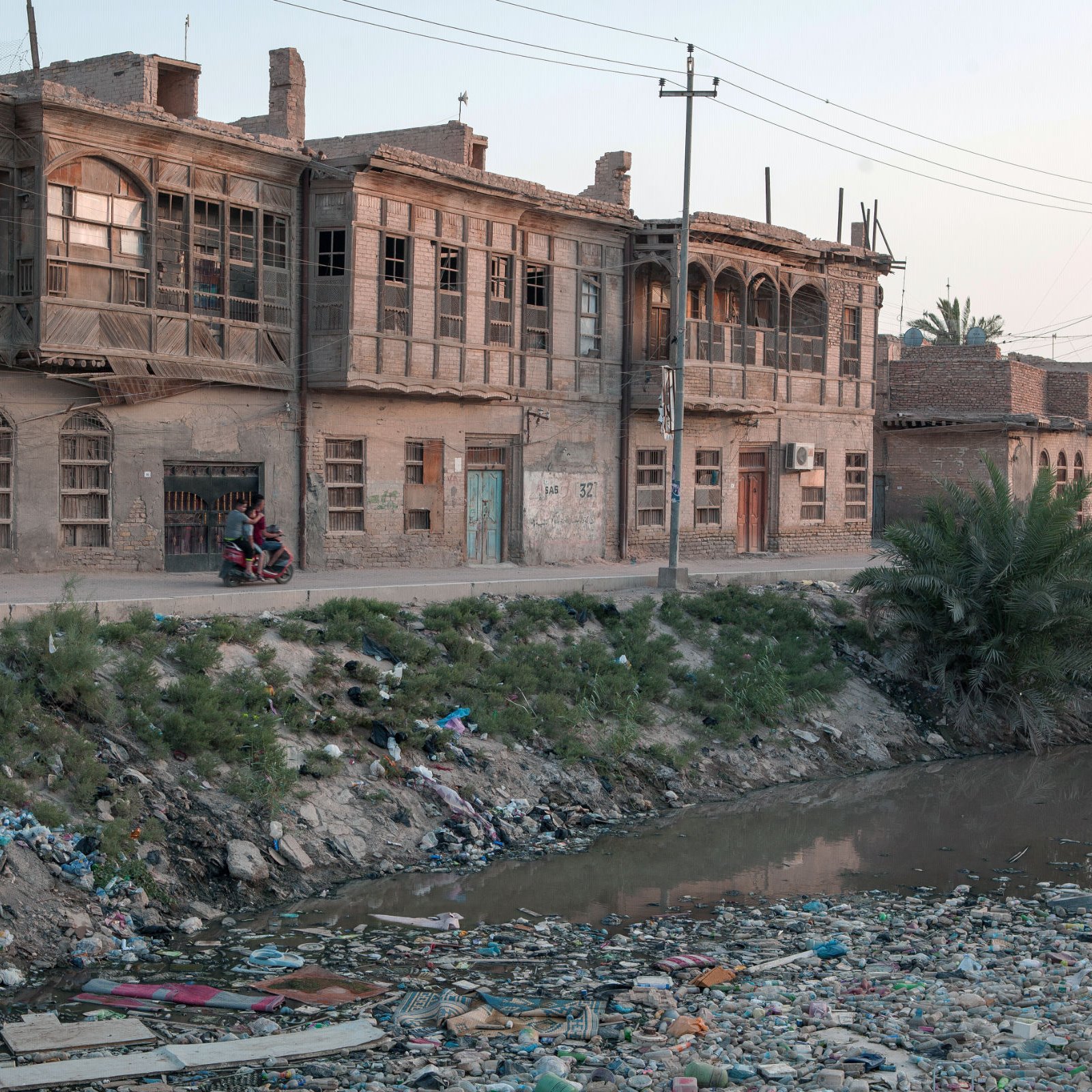 L'image montre une rue bordée de vieilles maisons en briques, caractérisées par des fenêtres en bois. À gauche, on aperçoit une voiture garée. Sur le côté droit, un groupe de personnes semble discuter. Au premier plan, un bras de rivière est visible, rempli de déchets et de débris, entouré de végétation. L'éclairage doux de la fin de journée crée une atmosphère nostalgique dans ce paysage urbain en dégradation.