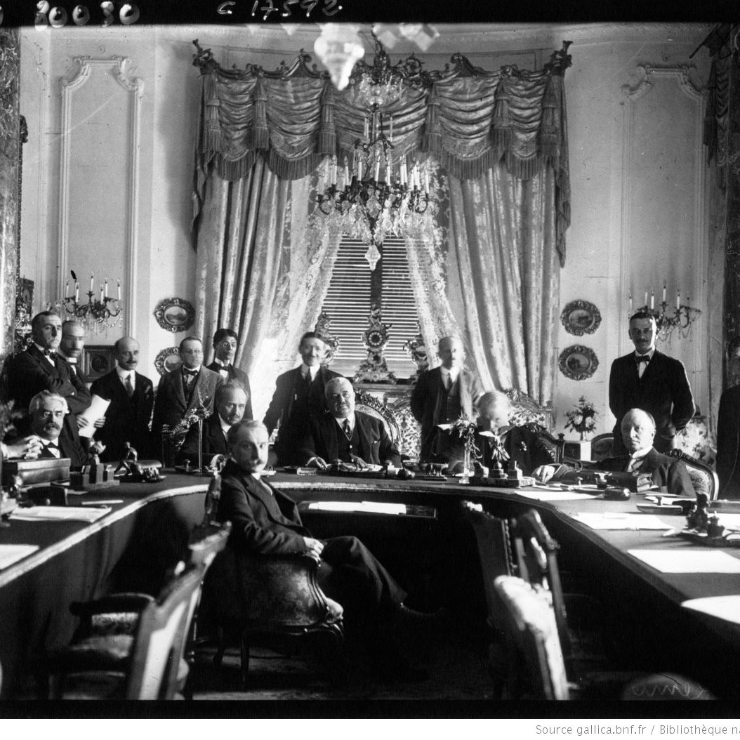 این تصویر، یک جلسه یا کنفرانس تاریخی را نشان می‌دهد که در آن گروهی از مردان در یک اتاق بزرگ و با طراحی تجملاتی گرد هم آمده‌اند. در مرکز تصویر، یک مردی با لباس رسمی نشسته است و دیگر افراد دور او ایستاده و نشسته‌اند. دکوراسیون اتاق شامل پرده‌های بلند، لوستر و تزئینات دیواری است که به فضای جلسه شکوه و جلال خاصی می‌بخشد. این تصویر نشان‌دهنده یک رویداد مهم و رسمی در تاریخ است.