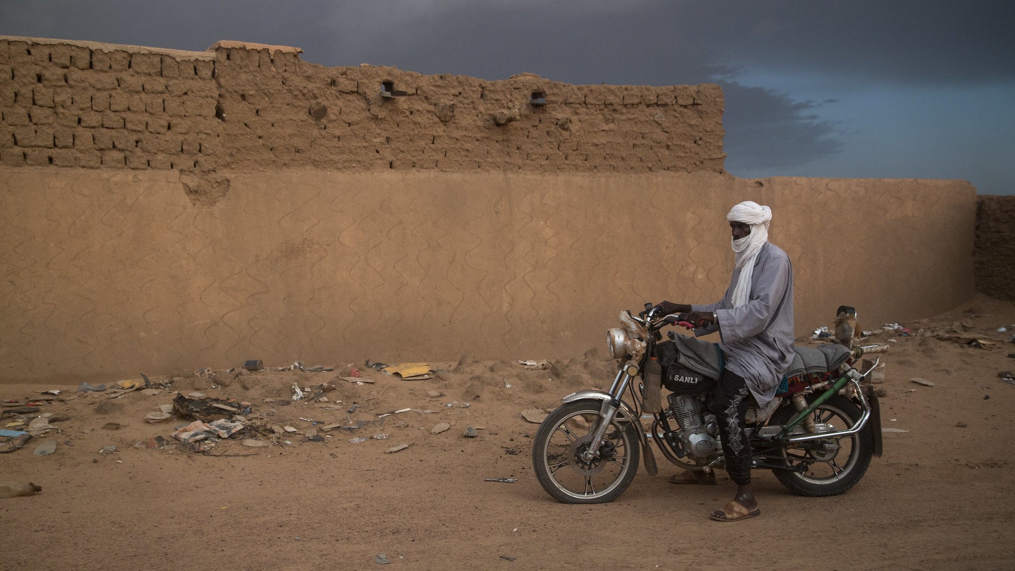 L'image montre un homme vêtu de vêtements traditionnels, avec un turban, debout à côté d'une moto. Il se trouve dans un environnement désertique, avec un mur de terre crue à l'arrière-plan. Le sol est poussiéreux et éparpillé de déchets. Le ciel semble nuageux, ajoutant une atmosphère dramatique à la scène.
