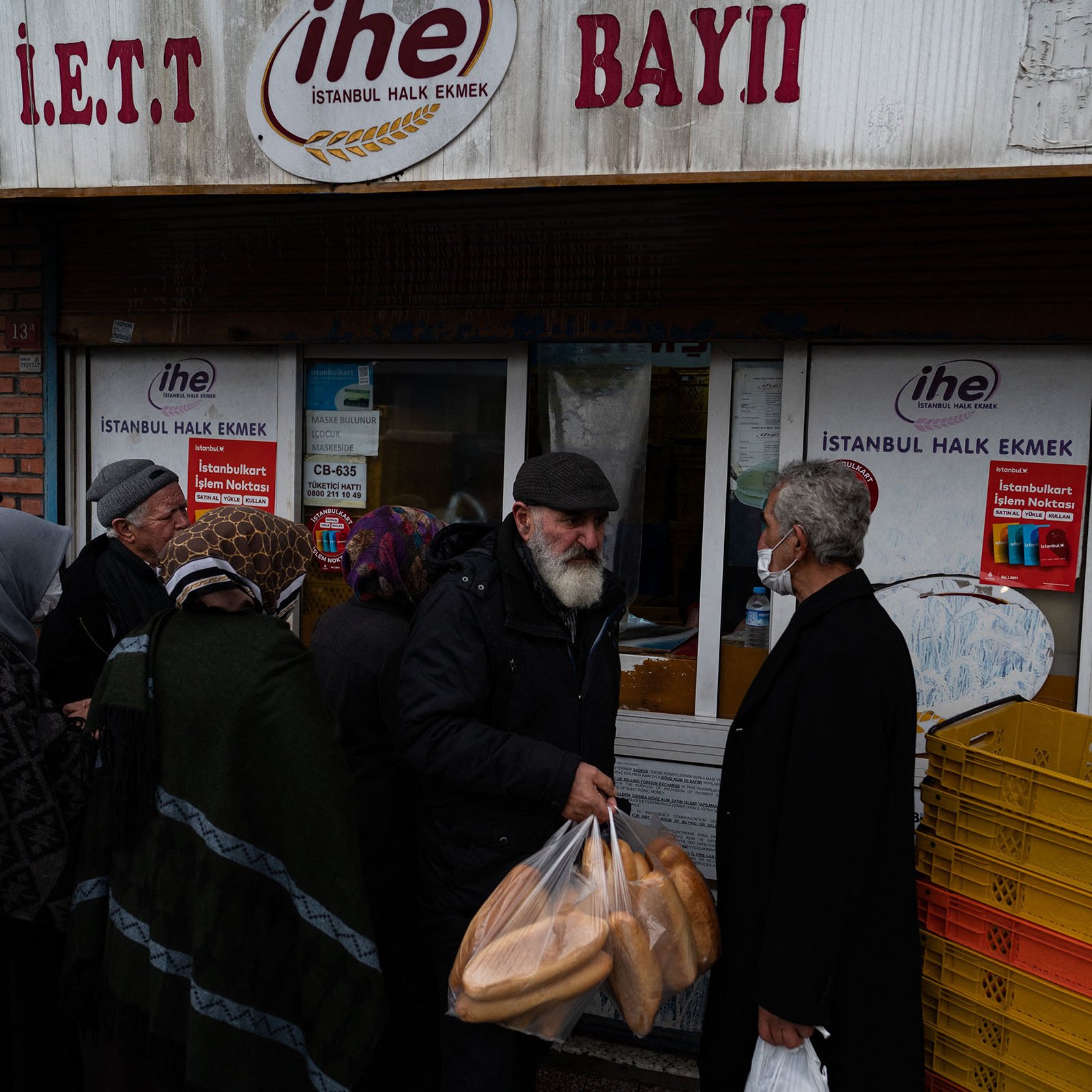 La imagen muestra a un grupo de personas en la calle, frente a una tienda. Algunos de ellos llevan bolsas en las manos, probablemente con pan. La escena sugiere una atmósfera de actividad comunitaria, ya que la gente está haciendo fila. Se pueden ver diferentes edades y estilos de ropa entre los presentes. Además, hay un letrero visible en la tienda que indica que se trata de un punto de venta de pan en Estambul.