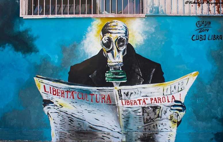 L'immagine rappresenta un murale che ritrae una figura umana con una maschera antigas, seduta e intenta a leggere un quotidiano. Il soggetto indossa un abbigliamento scuro e la scena è caratterizzata da una palette di colori vivaci, con sfumature di blu e bianco sullo sfondo. Le parole "LIBERTÀ", "CULTURA" e "LIBERTÀ di PAROLA!" sono scritte con un carattere forte e chiaro, sottolineando un messaggio di libertà e critica sociale. La data "2014" indica quando è stata realizzata l'opera, con il riferimento a "Cuba Libera" che suggerisce un legame con temi di libertà e diritti civili.