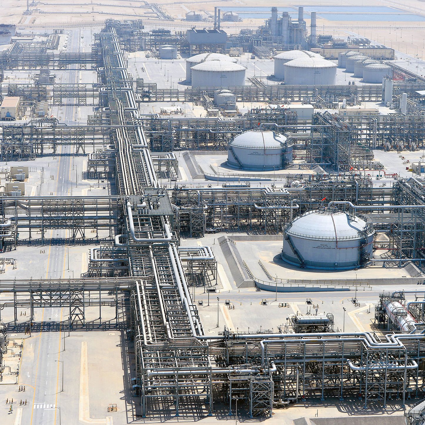 L'immagine mostra un impianto industriale, probabilmente una raffineria o un complesso di estrazione di prodotti petroliferi. Si possono vedere grandi serbatoi rotondi e una vasta rete di tubazioni che si snodano attraverso il sito. L'area circostante appare deserta, con una superficie di sabbia o terra arida. Sullo sfondo si intravedono strutture alte, che potrebbero essere torri di raffineria o impianti di trattamento. L'intero complesso è ben organizzato, con una disposizione sistematica delle tubazioni e dei serbatoi.