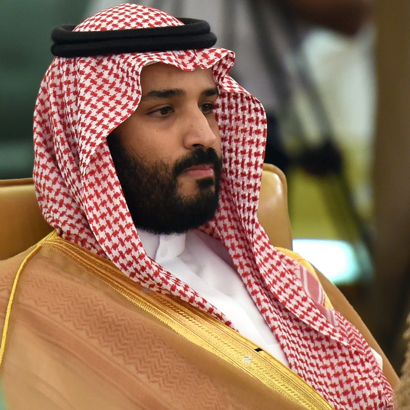 L'image montre un homme portant un long vêtement traditionnel arabe, composé d'une robe beige et d'un headdress appelé "ghutrah" ou "keffieh", qui est noué avec un bandeau noir. Il a une expression sérieuse et se trouve assis sur une chaise. En arrière-plan, des drapeaux peuvent être aperçus, suggérant un événement officiel ou une rencontre.