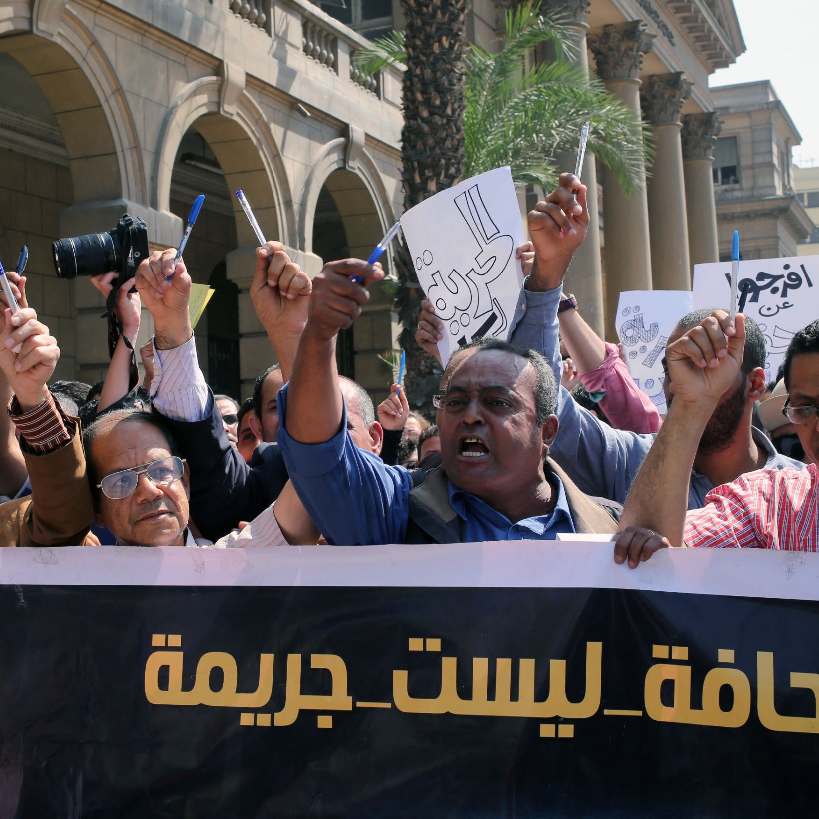 L'image montre une manifestation avec un groupe de personnes levant les bras et tenant des pancartes. Ils semblent exprimer leur mécontentement ou revendiquer des droits, notamment en rapport avec la liberté de la presse. L'atmosphère est énergique et déterminée, avec des visages montrant des émotions de passion et de revendication. À l'arrière-plan, on peut voir des bâtiments qui indiquent un environnement urbain. Le slogan sur la banderole semble défendre une position sur la presse, suggérant que celle-ci ne devrait pas être considérée comme un crime.