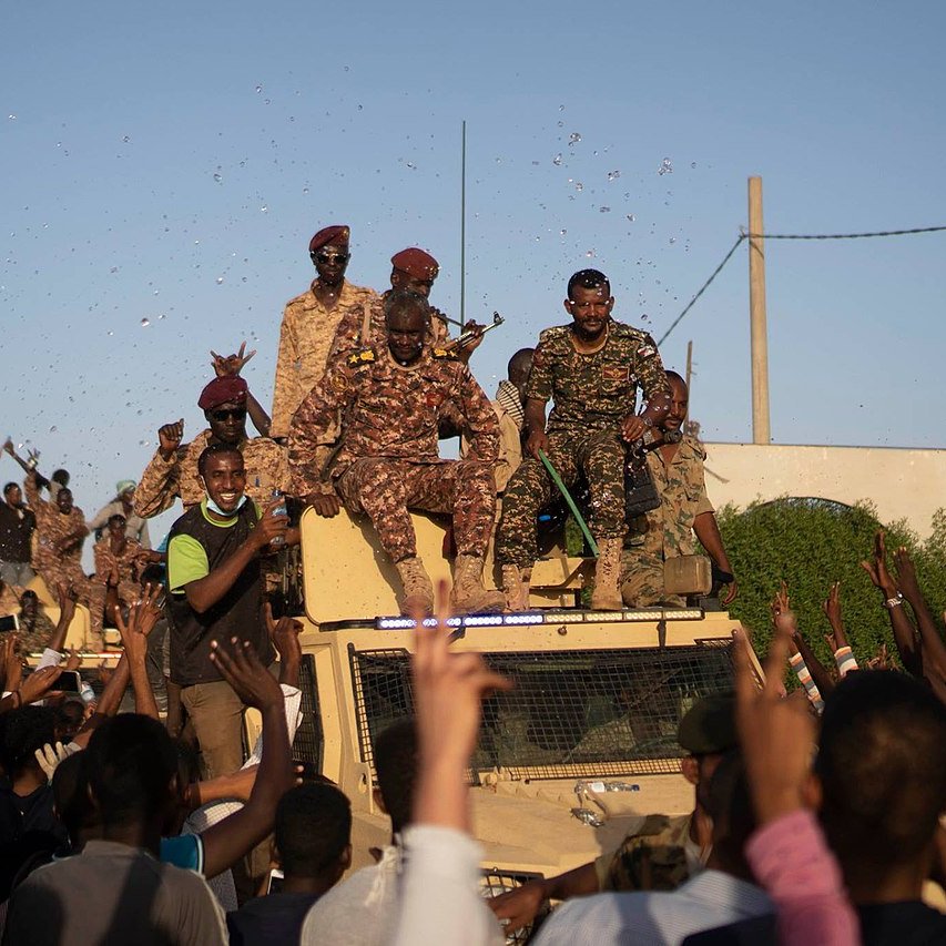 L'image montre un groupe de personnes en liesse rassemblées autour d'un véhicule militaire. Des soldats, vêtus d'uniformes camouflés, sont visibles en hauteur sur le véhicule, tandis que la foule en dessous lève les bras et semble manifester de la joie ou de l'enthousiasme. On peut apercevoir des confettis ou des éclats de couleur qui ajoutent à l'ambiance festive. L'atmosphère générale semble être celle d'une célébration ou d'un événement important.