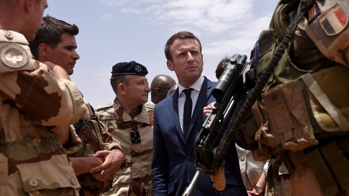 L'image montre un groupe de militaires en uniforme, dont certains portent des casques et des armes, rassemblés autour d'un homme en costume et cravate. Cet homme a une expression concentrée et semble en conversation avec les militaires, indiquant une interaction sérieuse. Le décor évoque un environnement militaire, probablement en extérieur, sous un ciel partiellement nuageux. Cette scène pourrait représenter une visite officielle ou une interaction avec des forces armées.
