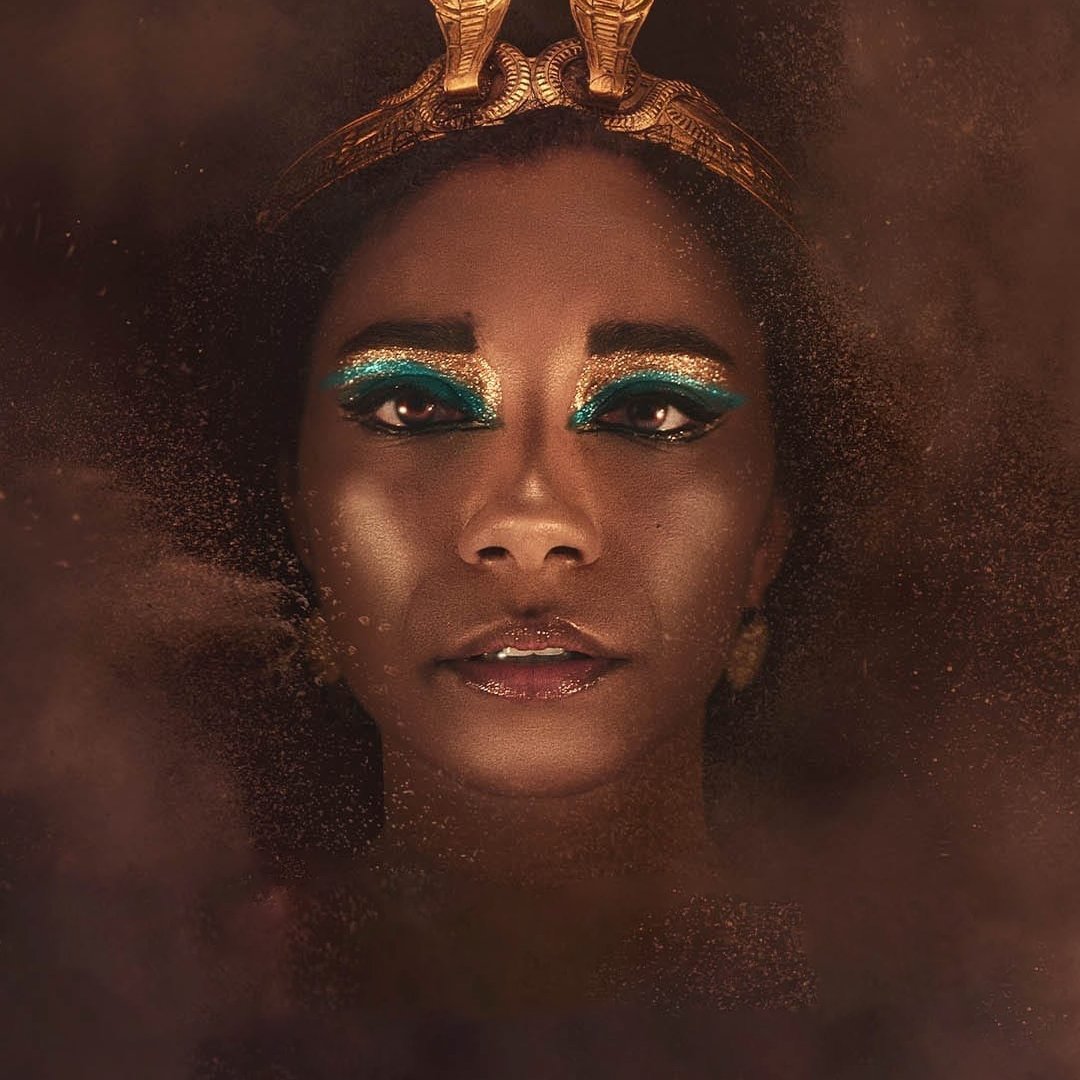 L'image montre une personne au visage ornée, avec un maquillage très artistique mettant en avant des yeux accentués par une ombre à paupières bleu vif. La personne porte une coiffe dorée en forme de serpents, ajoutant à l'aspect majestueux de l'ensemble. Le fond est flou et semble composé de particules dorées, créant une atmosphère mystique et intrigante. L'expression du visage est à la fois sereine et mystérieuse.