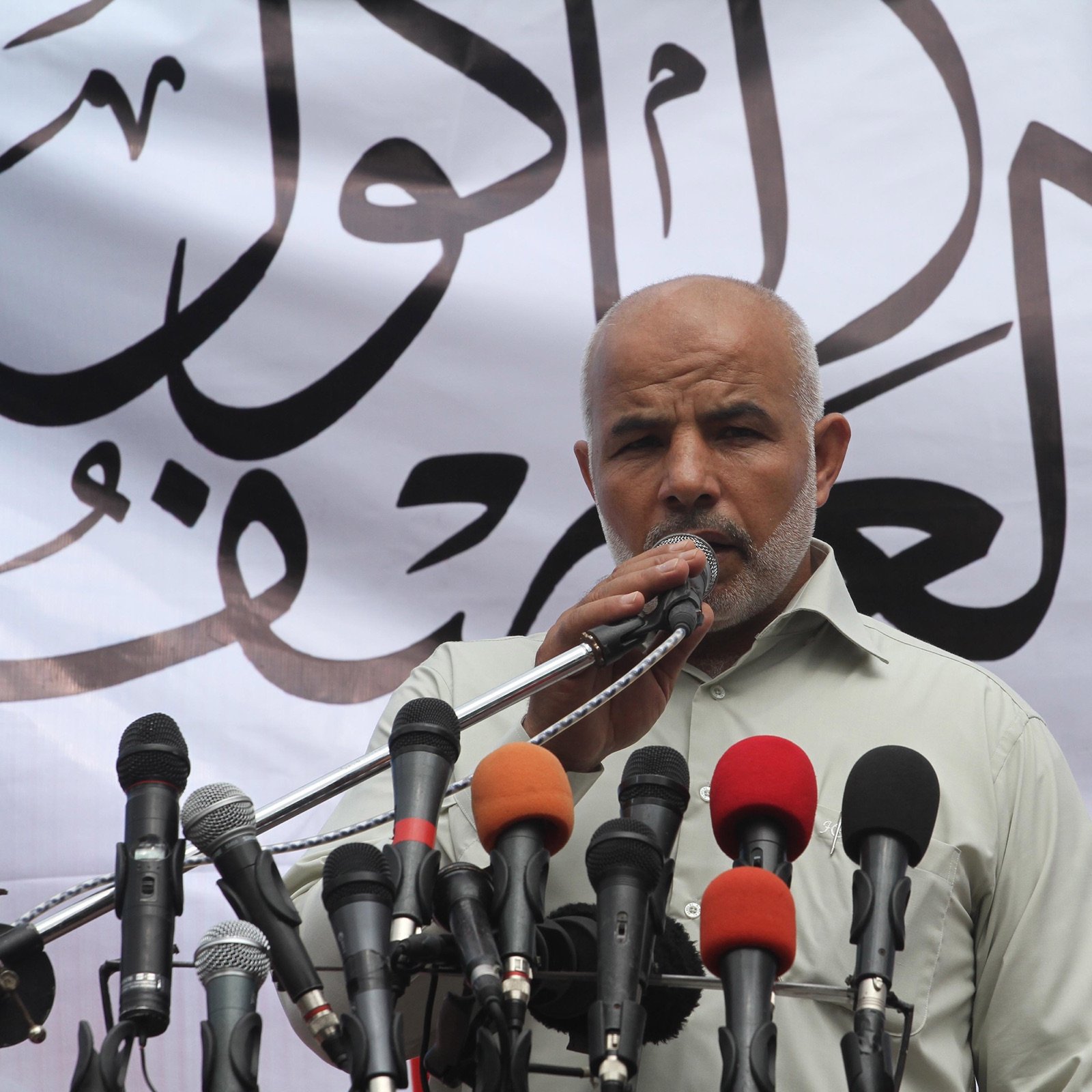 L'image montre un homme debout devant un microphone, prenant la parole lors d'un événement. Il a une barbe grise et porte une chemise claire. Derrière lui, un grand panneau blanc est décoré d'écritures arabes. Sur le devant, plusieurs microphones de différentes couleurs sont alignés, ce qui indique qu'il s'agit probablement d'une conférence de presse ou d'un discours public. L'atmosphère semble solennelle et engagée.