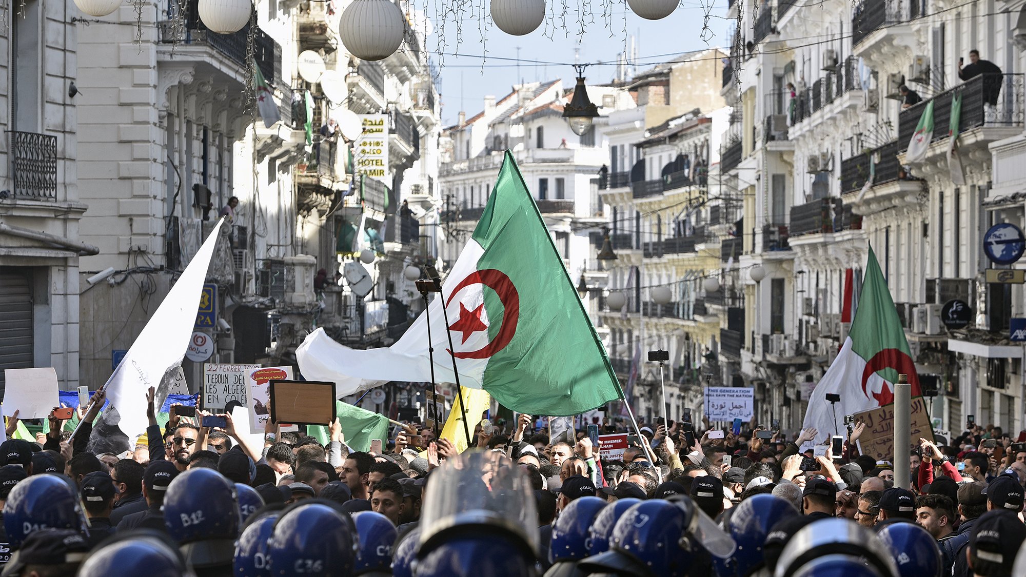 تظهر الصورة تجمعًا حاشدًا في شارع بالجزائر، حيث يحمل المتظاهرون الأعلام الجزائرية ويعبرون عن مطالبهم. يوجد في مقدمة الصورة رجال شرطة يرتدون خوذات واقية، وكأنهم يراقبون الوضع. يُلاحظ وجود لافتات ومحتجين يعبرون عن آراءهم، مما يدل على حماس واهتمام المواطنين بالقضايا المحلية. الأجواء تبدو مليئة بالحيوية والتوتر في آن واحد.