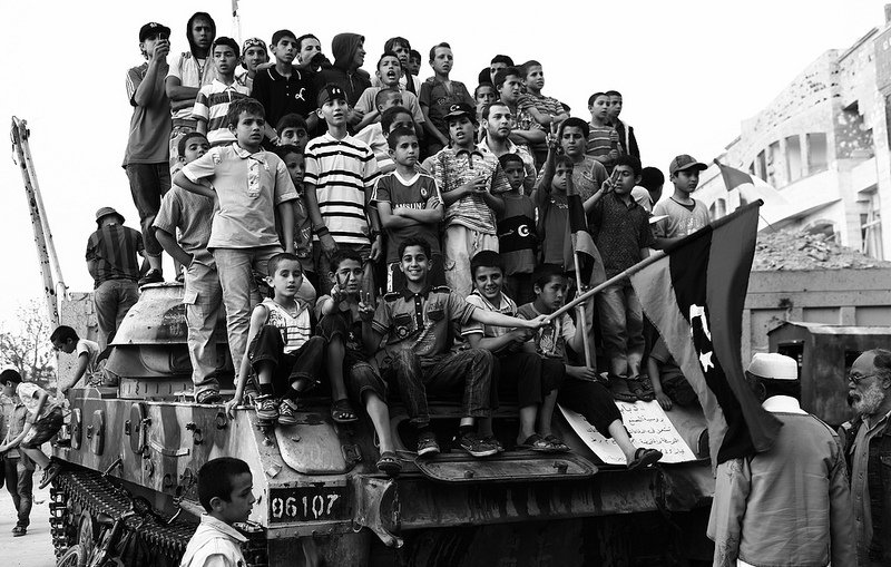 L'image montre un grand groupe d'enfants rassemblés sur un char, probablement dans un contexte de célébration ou de manifestation. Les enfants affichent diverses expressions, allant de la joie à la curiosité. Ils portent des vêtements colorés, souvent rayés, et certains tiennent un drapeau. En arrière-plan, on peut voir des bâtiments et des ruines, suggérant un environnement marqué par des conflits ou des événements historiques. L'image est en noir et blanc, ce qui accentue le côté dramatique de la scène.