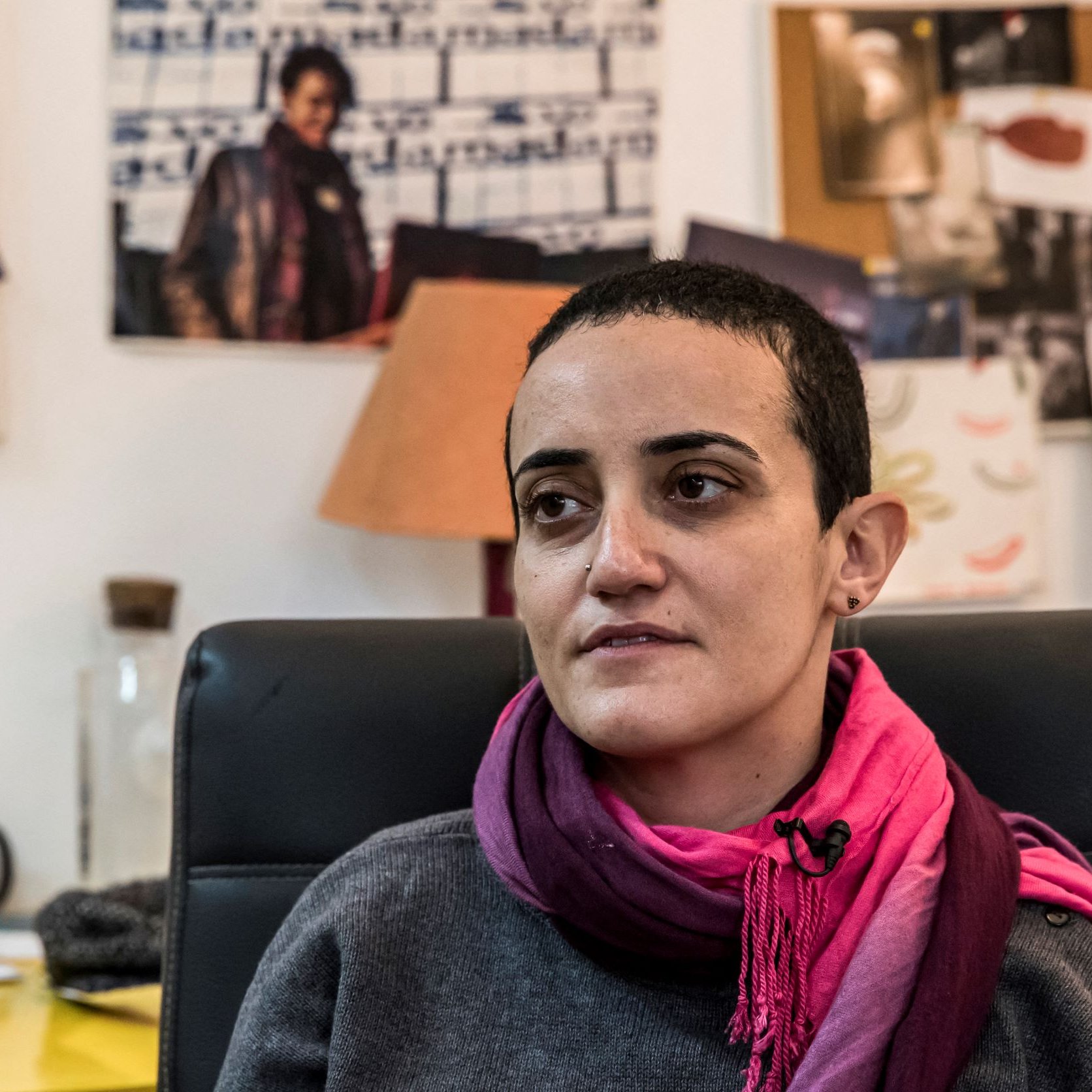 تظهر الصورة امرأة في مكتب، حيث تضع على كتفيها وشاحًا ورديًا. تصفح عينيها بعمق، ويبدو أنها تفكر أو تعبر عن مشاعر معينة. في الخلفية، يمكن رؤية مجموعة من الأوراق والصور المعلقة على الحائط، بالإضافة إلى أضواء وزرع، مما يعطي انطباعًا عن بيئة العمل أو الإبداع. الأجواء تبدو غير رسمية، وتعكس شخصية المرأة المتواجدة في الصورة.