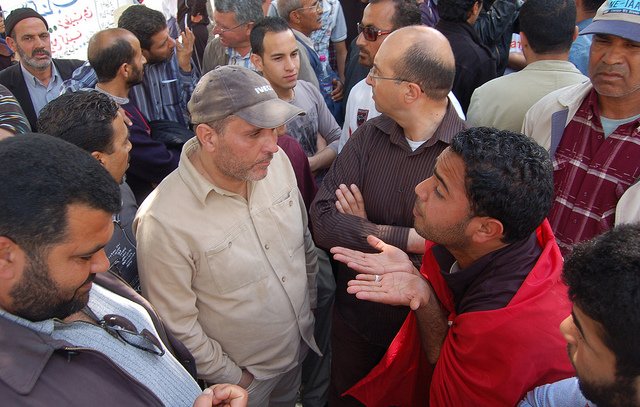 تظهر الصورة مجموعة من الرجال مجتمعين في مكان واحد، يبدو عليهم الحماس والنقاش. هناك شخص يرتدي قميصاً بلون فاتح يتحدث بجدية مع شخص آخر يرتدي سترة داكنة. بعض الرجال الآخرين يتابعون الحوار، ويظهر بينهم تعبيرات مختلفة تعكس تفاعلهم مع النقاش. الخلفية تحتوي على مجموعة من الأشخاص الآخرين، مما يوحي بأن المكان مزدحم ومليء بالحيوية. يبدو أن الوضع يعكس تجمعاً اجتماعياً أو نقاشاً حيوياً حول قضية معينة.