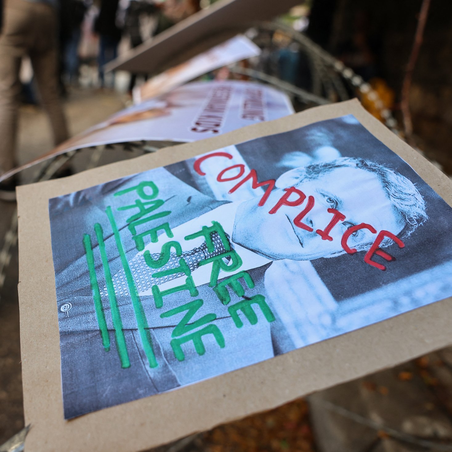 L'immagine mostra un cartello posizionato su una recinzione, con il volto di una persona ritratta in bianco e nero. Sotto l'immagine ci sono delle parole scritte con pennarelli colorati: "COMPLICE" e "FREE PALESTINE". Il contesto sembra essere un evento di protesta, con altri cartelli visibili in background e una folla di persone. L'atmosfera è quella di un'espressione di dissenso e di richiesta di libertà per la Palestina.