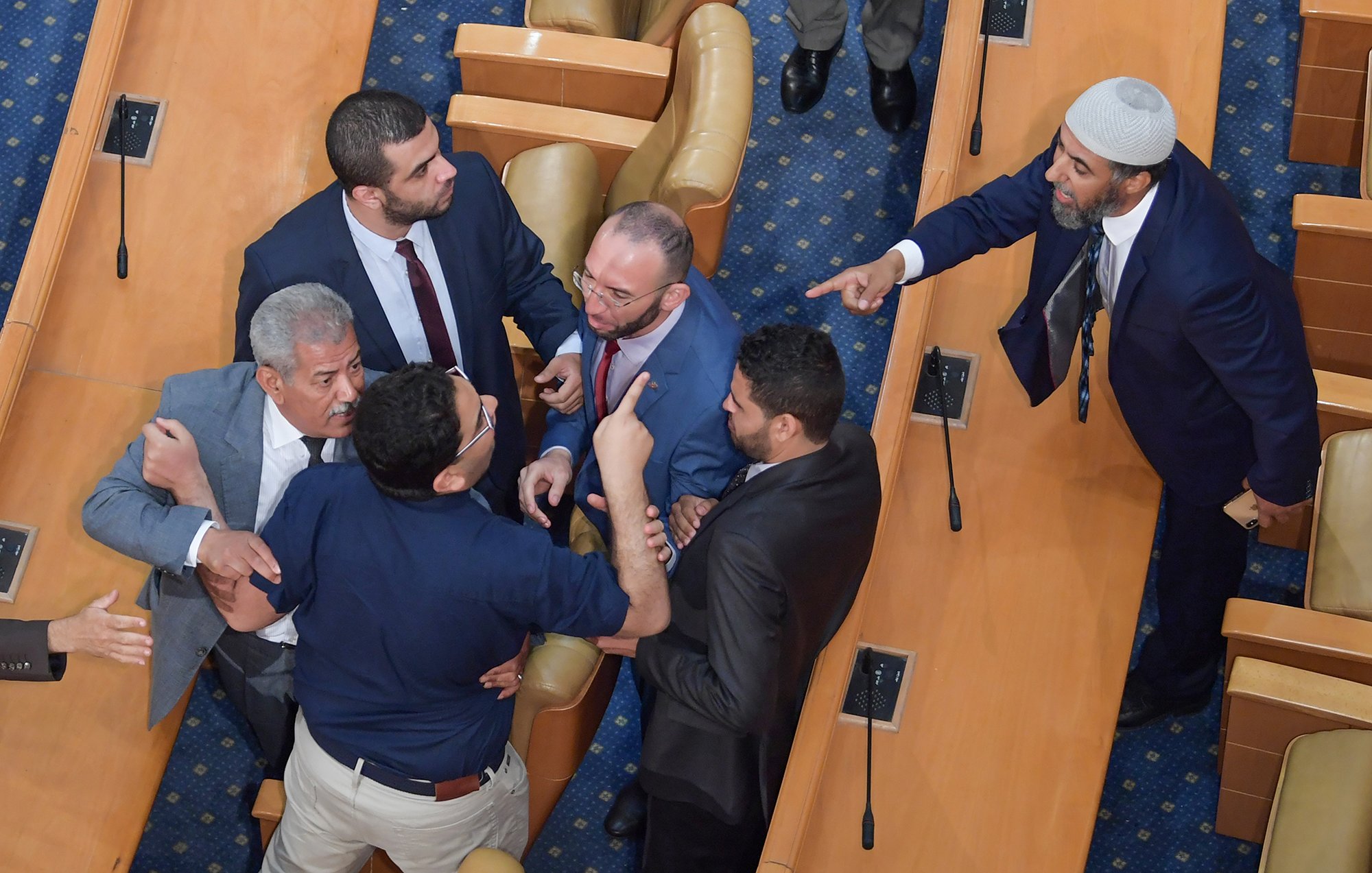 L'image montre une scène tendue dans une assemblée ou un parlement. Plusieurs hommes discutent vivement, certains pointant du doigt et affichant des expressions agitées. Il y a une mise en scène de désaccord, avec des gestes éloquents et une proximité physique entre les participants. Le décor est simple, avec des sièges en bois et un tapis bleu au sol, suggérant un environnement formel.
