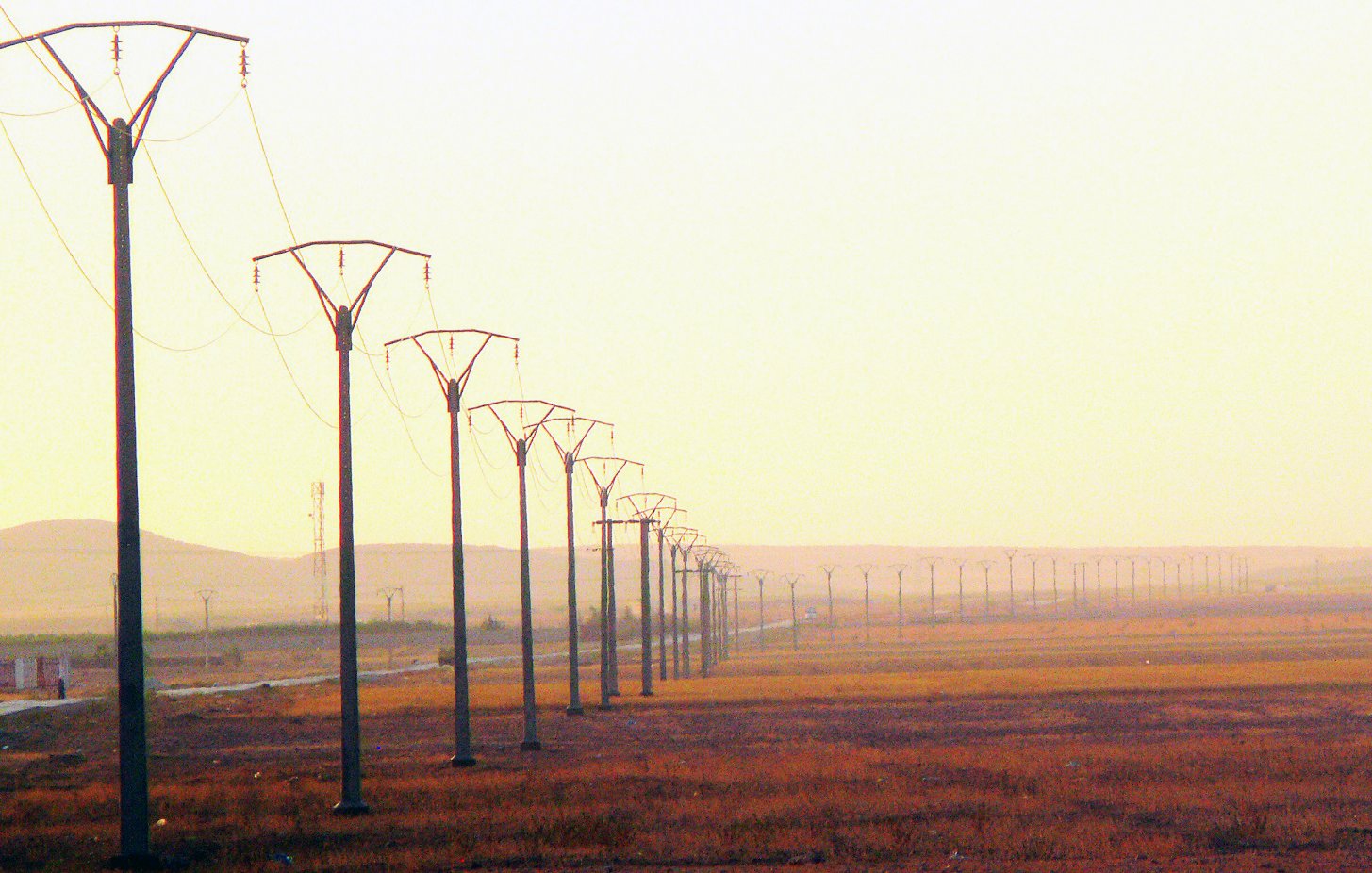 L'image montre une perspective sur un paysage de campagne, dominé par des pylônes électriques s'étendant sur une longue distance. Le sol est principalement sec et herbeux, avec des teintes dorées, suggérant la lumière douce d'un lever ou d'un coucher de soleil. En arrière-plan, on peut apercevoir des collines ou des montagnes floues, contribuant à une ambiance calme et désertique. L'ensemble évoque un sentiment d'immensité et de tranquillité.