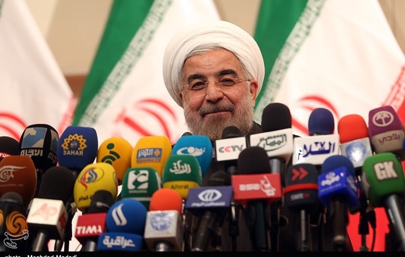 L'image montre un homme souriant, vêtu d'une tenue formelle et portant un turban blanc. Il se tient devant une multitude de microphones de différentes chaînes de télévision et médias. En arrière-plan, il y a des drapeaux avec des motifs et des couleurs qui semblent représenter un pays. L'ambiance semble être celle d'une conférence de presse ou d'un événement médiatique.