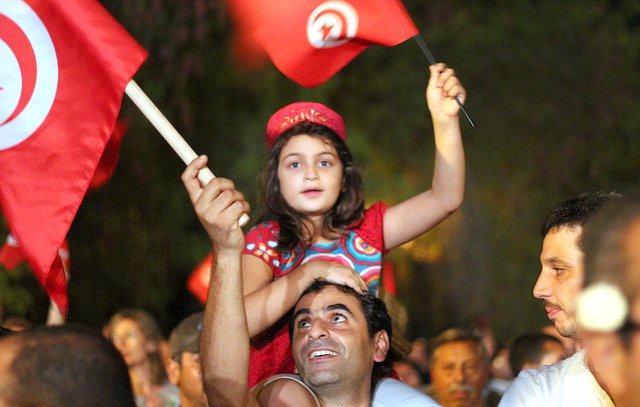 L'image montre une scène de célébration en Tunisie. Une jeune fille, vêtue d'une robe colorée, est assise sur les épaules d'un homme qui sourit. Ils brandissent des drapeaux tunisiens. En arrière-plan, on aperçoit une foule joyeuse, créant une atmosphère festive. Les expressions des visages reflètent la joie et l'engagement.