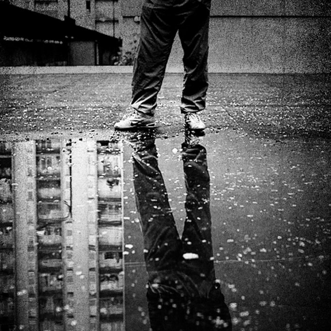 تظهر في هذه الصورة مجموعة من العناصر المثيرة للاهتمام. في المقدمة، نقف أمام شخص يرتدي بنطالًا و حذاء، واقفًا فوق سطح مغطى بالماء المتجمع، مما يشكل بركة صغيرة. يظهر انعكاس الشخص في الماء، مما يضيف بعدًا جماليًا للصورة. الخلفية عبارة عن جدار رمادي وسطح فارغ، مما يساعد على تركيز الانتباه على الشخص والانعكاس في الماء. الجو العام يبدو هادئًا ومظلمًا، مع تباين قوي بين الألوان.