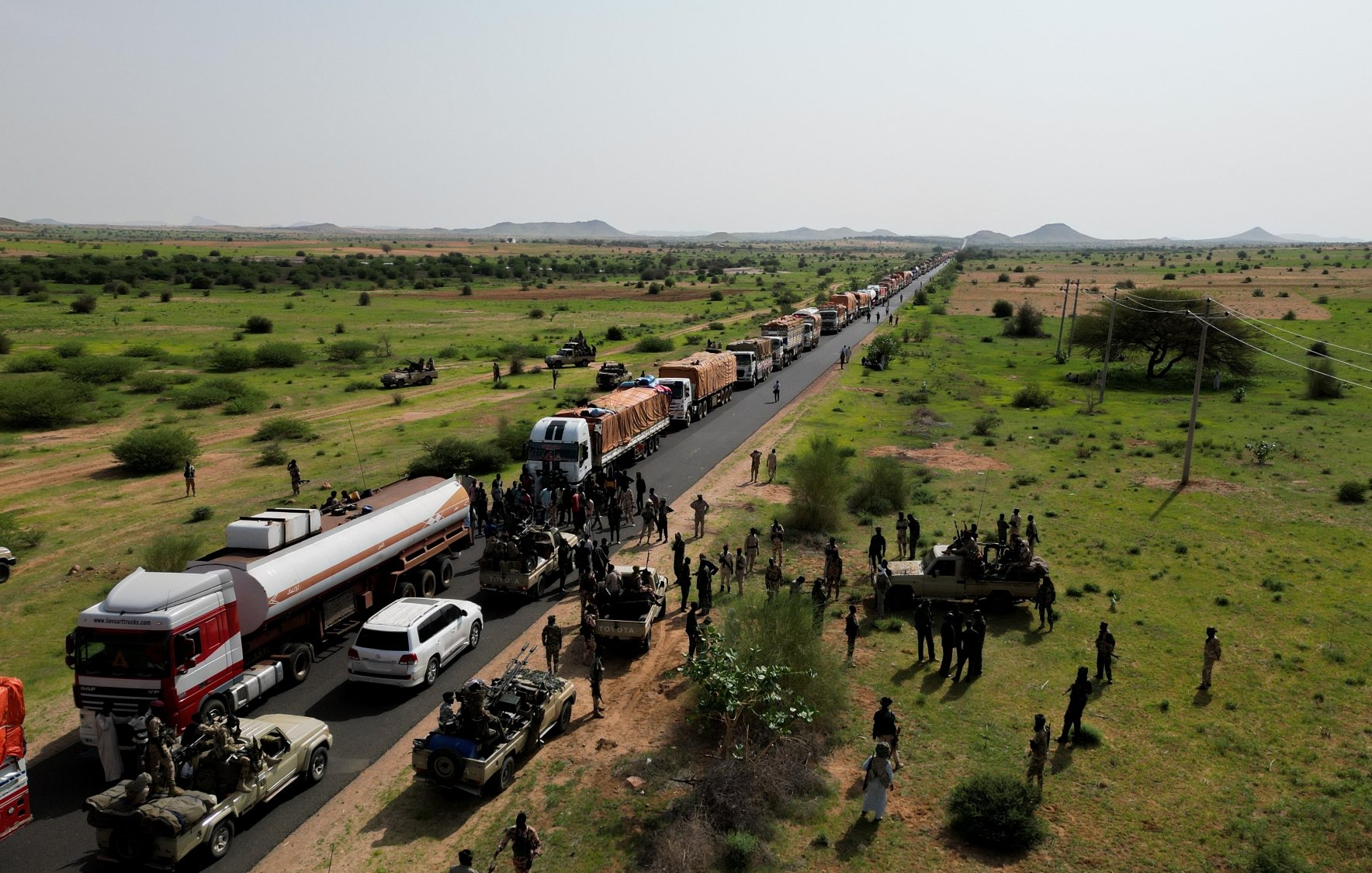 Convoi de camions sur une route, entouré de militaires et de verdure désertique.
