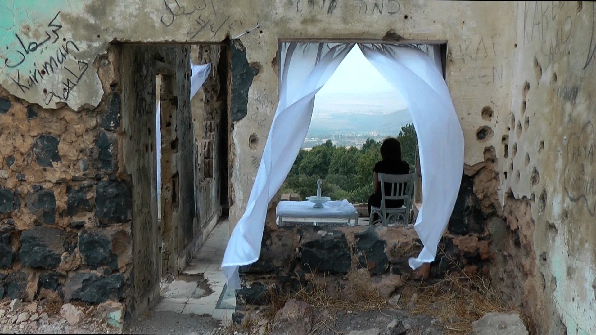 L'image montre l'intérieur d'une vieille maison en ruines, dont les murs sont en pierre et présentent des signes de dégradation avec des inscriptions. À travers une ouverture, on peut voir un paysage verdoyant au loin. Une chaise est placée à l'intérieur, avec une table dressée devant elle, complétée par des rideaux blancs qui flottent doucement. À côté de la table, une silhouette assise, probablement une personne contemplant le paysage. L'atmosphère générale de la scène évoque la mélancolie et la tranquillité.