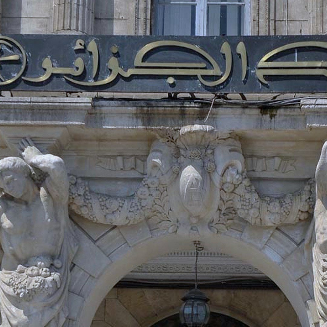 La imagen muestra un edificio con una arquitectura clásica. En la parte superior, hay un letrero que dice "بنك الجزائر" (Banco de Argelia). A ambos lados del acceso principal, se pueden ver esculturas de figuras masculinas sosteniendo lo que parecen ser cestas de frutas. La fachada del edificio tiene detalles ornamentales elaborados y columnas, lo que le da un aspecto majestuoso.