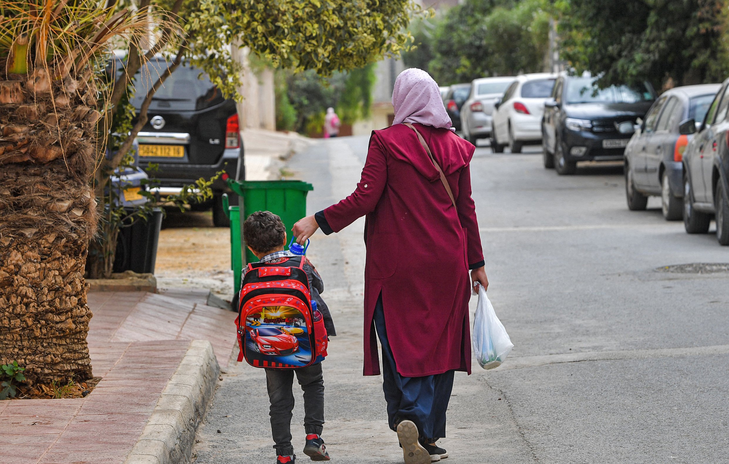 The image shows a woman walking down a street with a young child. The woman is wearing a long maroon coat and a headscarf, while the child has a colorful backpack with a cartoon character design. They are walking hand in hand along a road lined with parked cars and greenery on the sides. The atmosphere appears casual and everyday, suggesting they are possibly returning from school or heading to a local destination.