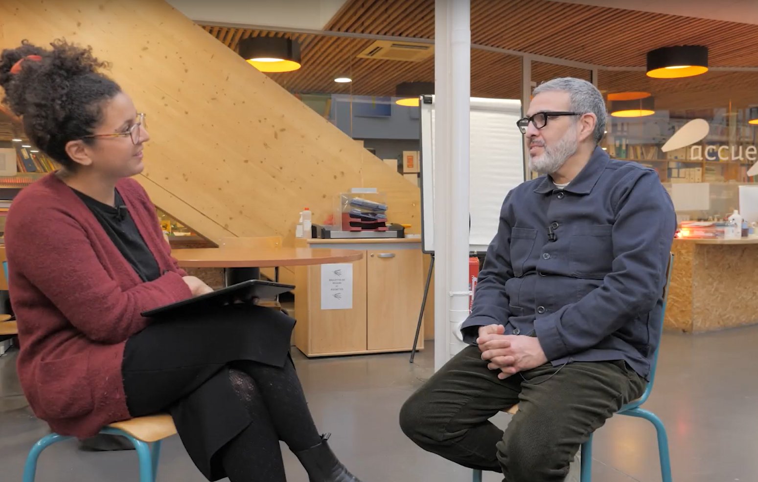 L'image montre deux personnes assises dans un espace intérieur. À gauche, une femme avec des cheveux bouclés et des lunettes, vêtue d'un pull rouge, tient une tablette. À droite, un homme portant des lunettes et une chemise bleu foncé est assis, les mains sur ses genoux. L'environnement est moderne, avec des murs en bois et des éléments de décoration qui donnent une ambiance chaleureuse. Cela semble être une interview ou une discussion.