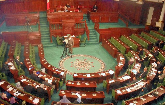 L'image montre l'intérieur d'un parlement, probablement un hémicycle de l'assemblée nationale. On peut voir plusieurs rangées de sièges verts disposés en arc de cercle, où des membres semblent assister à une session. Au centre, un tapis décoratif est visible, et à l'arrière-plan, on aperçoit une estrade pour les orateurs. Des caméras sont présentes, indiquant que la session est probablement diffusée. L'ensemble dégage une atmosphère formelle et politique.