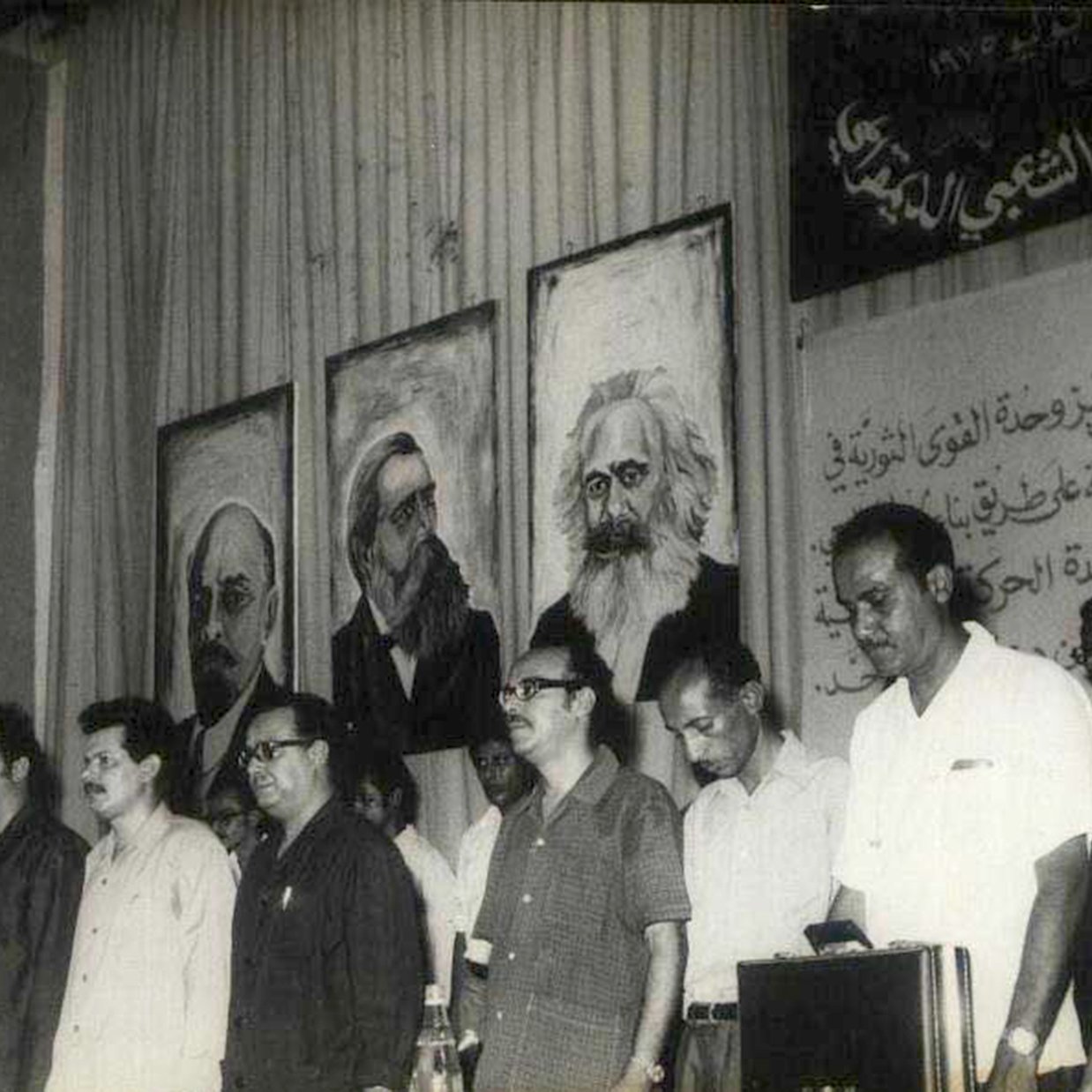 این تصویر مربوط به یک تجمع یا کنفرانس سیاسی به نظر می‌رسد. در پس‌زمینه، تابلوهایی از چهره‌های معروف تاریخی یا سیاسی وجود دارد. افرادی که در جلوی صحنه ایستاده‌اند، موضع جدی و متفکرانه‌ای دارند و در حال گوش دادن به سخنرانی یا برنامه‌ای هستند. فضای کلی تصویر به نظر می‌رسد که تمرکز بر مسائل اجتماعی و سیاسی داشته باشد. دیوار پشت‌شان نیز با متنی به زبان عربی تزئین شده است.