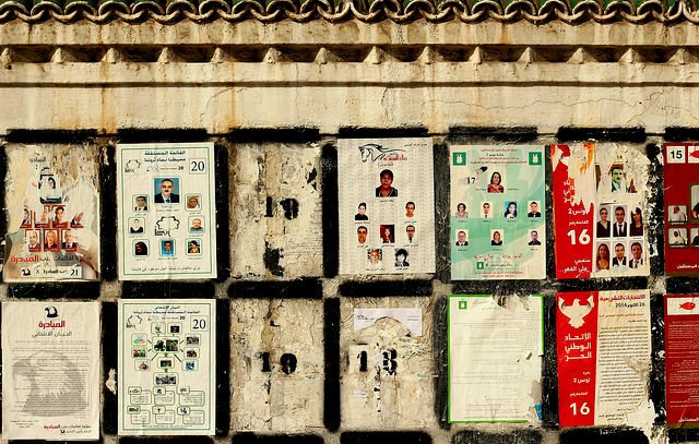 L'image montre un mur sur lequel sont affichés plusieurs documents et affiches. Ces derniers semblent être en lien avec des élections, avec des plaques présentant des photos et des noms de candidats. Certaines affiches sont abîmées et décolorées, révélant un certain vieillissement et une usure du temps. Le mur lui-même présente une texture rugueuse et des motifs décoratifs en haut. Les divers éléments placardés créent une composition visuelle chargée et informative.