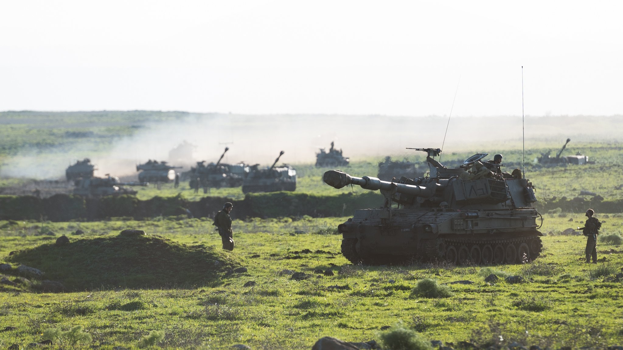 L'image montre un paysage militaire verdoyant avec des chars d'assaut et d'autres véhicules en arrière-plan. On peut voir un char principal en premier plan, déployé sur un terrain herbeux, tandis que plusieurs autres véhicules sont visibles plus loin, entourés d'une légère brume ou fumée. Quelques soldats sont présents, donnant une impression d'activité militaire dans un environnement naturel. La lumière est douce, suggérant peut-être un moment du jour comme le lever ou le coucher du soleil.