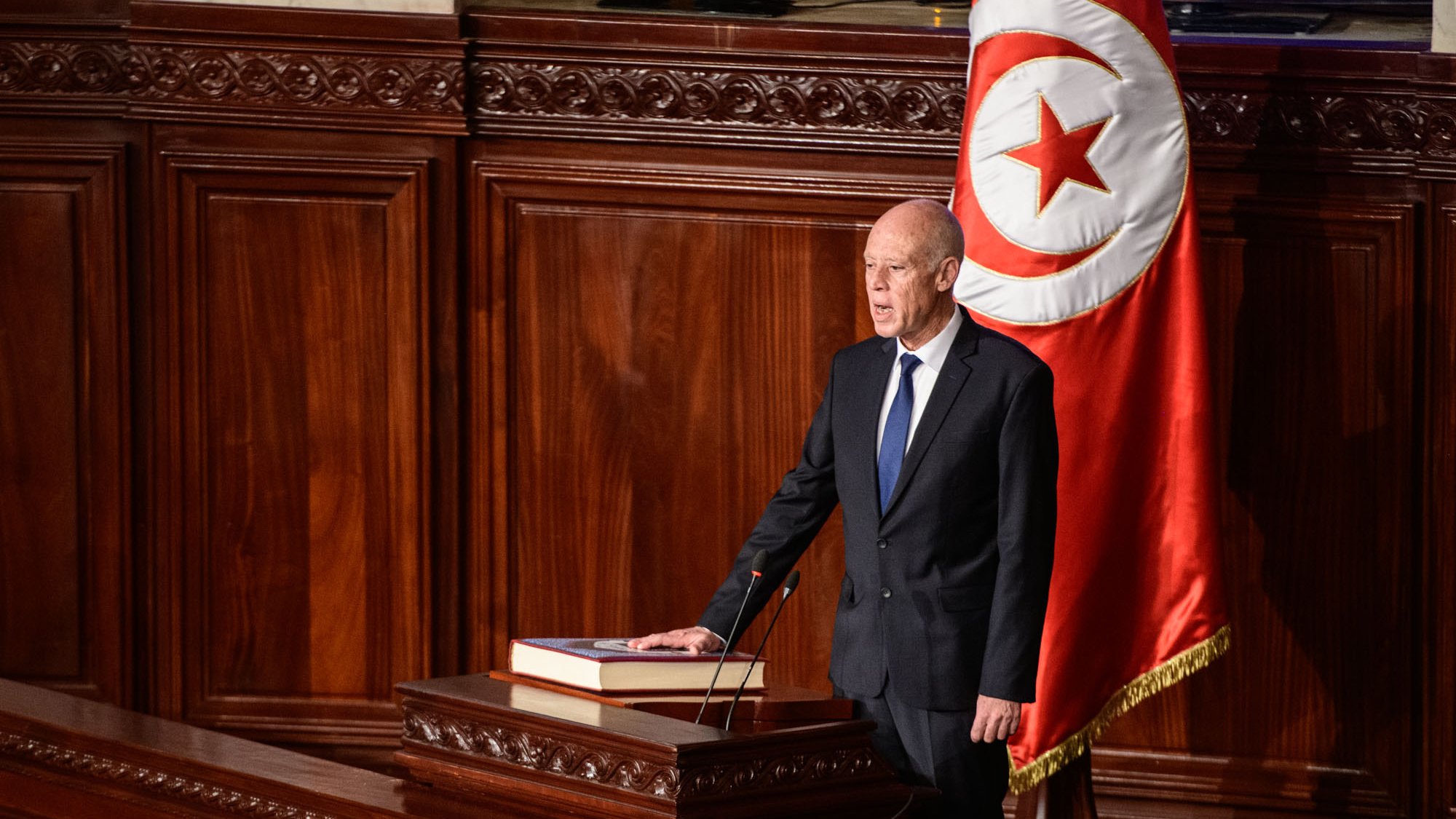 The image depicts a formal setting, likely in a government assembly or parliament. A man, dressed in a dark suit and tie, stands at a podium with a large book placed on it. Behind him, there is a prominent flag featuring a red background with a white circle and a red crescent and star, indicating it is Tunisia's national flag. The environment appears ornate, with wooden paneling and an overall serious ambiance, suggesting that he is addressing a legislative body or delivering an important speech.
