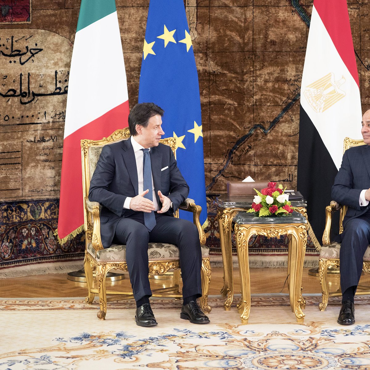 در این تصویر دو فرد در حال گفت‌وگو هستند. هر دو نفر در نشیمنگاه‌های مجلل و طلایی نشسته‌اند. در پس‌زمینه، پرچم‌های ایتالیا و مصر دیده می‌شوند. همچنین، پرچم اتحادیه اروپا نیز در تصویر وجود دارد. فضایی رسمی و دیپلماتیک حاکم است و گل‌های تزئینی روی میز بین دو نفر قرار دارد. این تصویر به نظر می‌رسد که نماای از یک دیدار یا مذاکرات رسمی باشد.