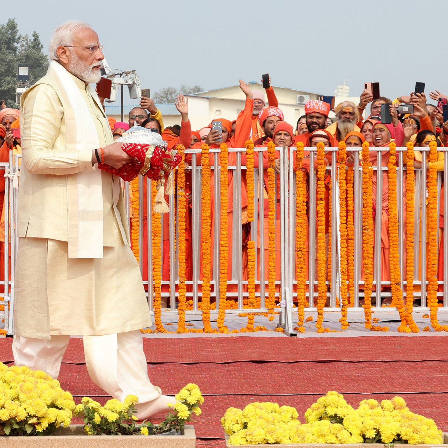 En la imagen se puede ver a un hombre con barba y cabello canoso vestido con un traje tradicional claro, que sostiene un objeto en sus manos. Este hombre camina hacia la derecha, mientras que detrás de él hay un grupo de personas vistiendo túnicas naranjas. Están emocionadas y algunas de ellas levantan sus teléfonos móviles para capturar el momento. La escena está decorada con flores amarillas y hay una valla blanca que separa al hombre de la multitud. En el fondo, se observa un ambiente festivo y colorido.